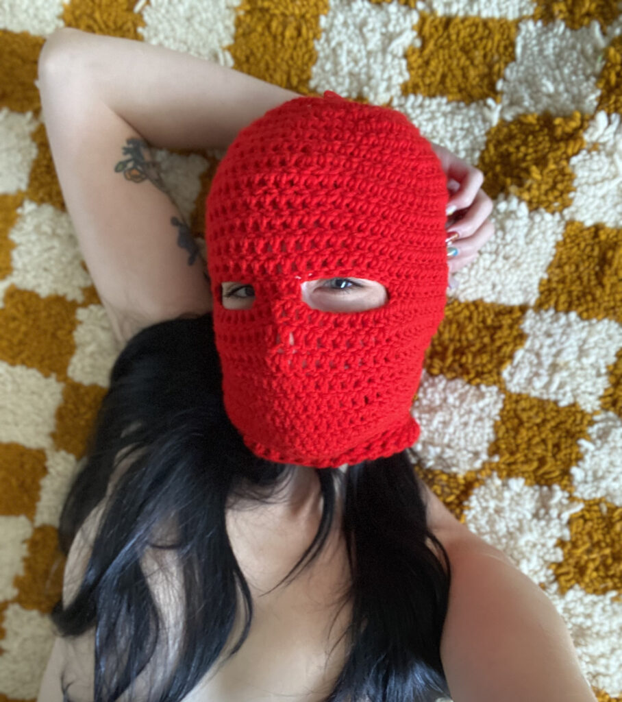 A selfie of me in a red crocheted balaclava, laying down on a cream and mustard-colored checkered rug