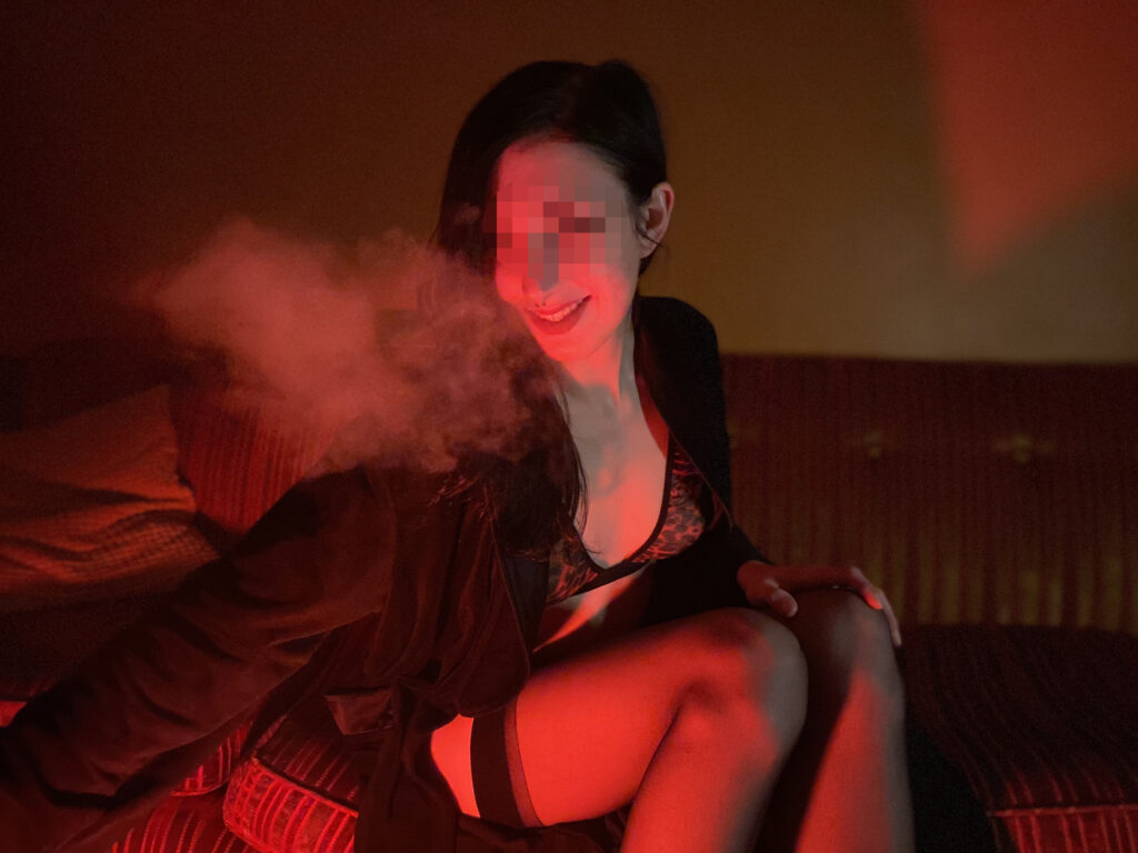 A photo of me leaning forward as I exhale smoke, sitting on a couch. A red light illuminates my face and side of my body. I'm wearing a red cheetah print lingerie set and a black robe.