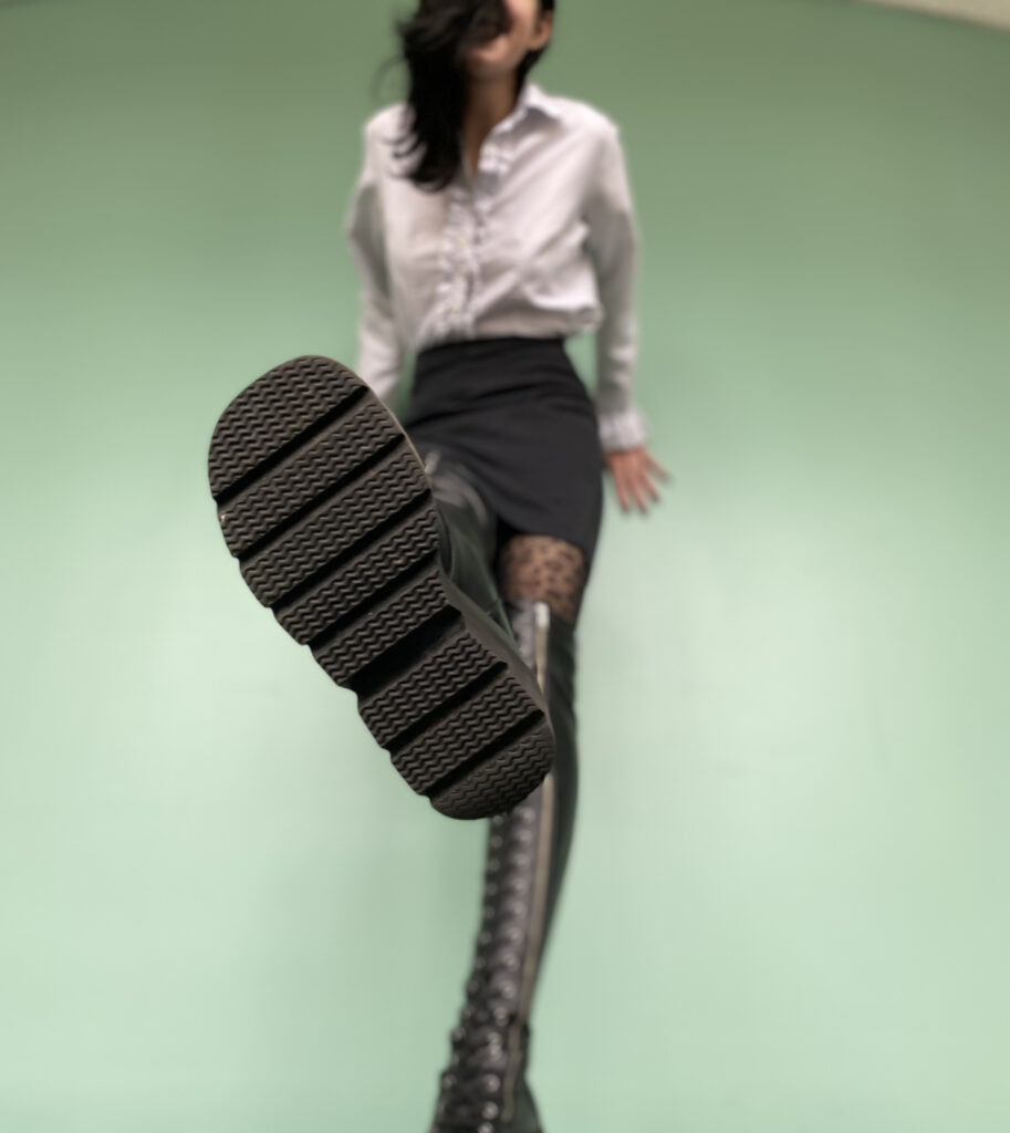 A photo from a lower perspective of me in a slutty business-casual outfit and thigh high boots. I'm lifting one leg and putting the sole of my shoe in front of the camera, as if to stomp on it. The background is out of focus.