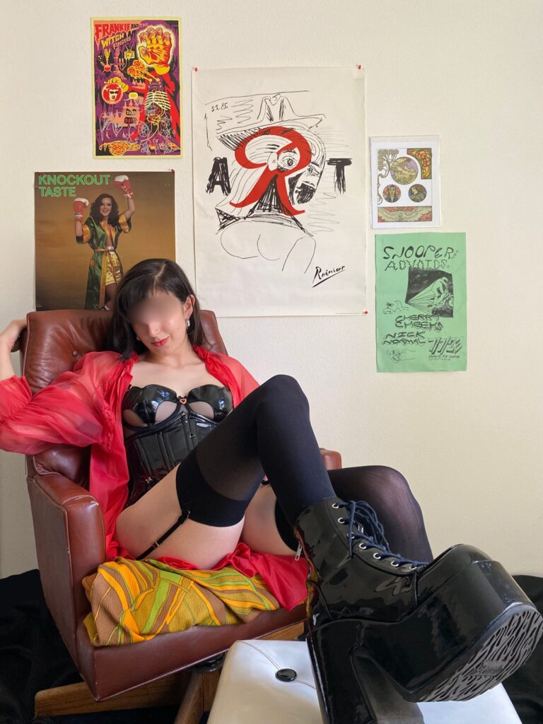 A photo of me in a black pvc bra, panty, and garter set and an open red nightgown. Im wearing sheer black stockings and ankle height platform pvc boots. I'm reclining in a vintage leather office chair and kicking my boots up on an ottoman