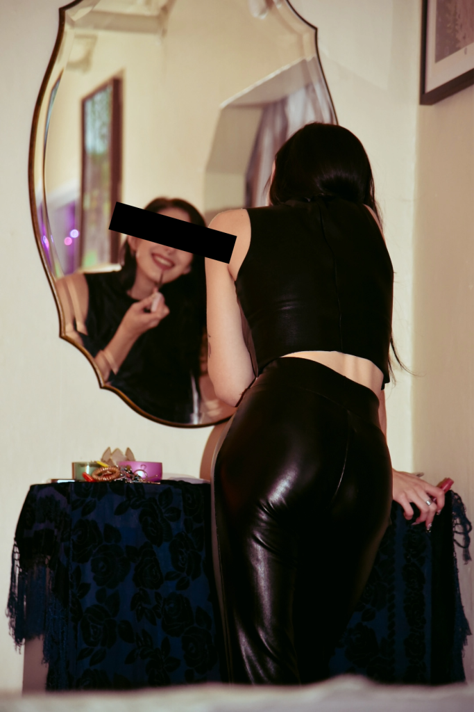 A photo of me from behind, smiling while I fix my lipstick in a vanity mirror. I'm wearing a black vinyl tank top and leggings